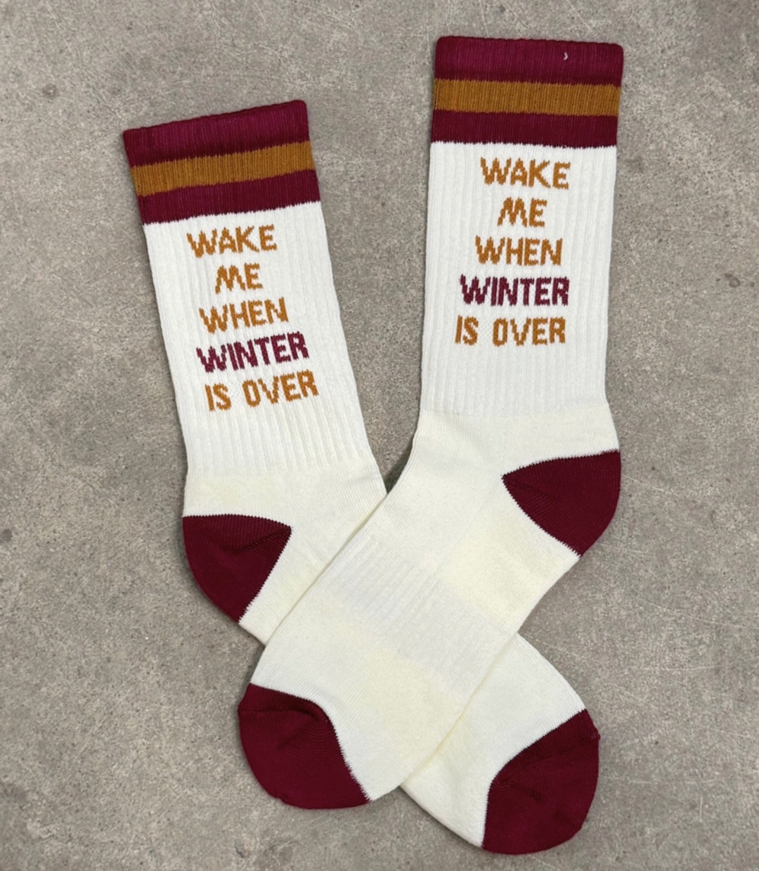 A pair of cream unisex socks with a text pattern that reads 'WAKE ME WHEN WINTER IS OVER' in a playful font, and a maroon heel and brown cuff.